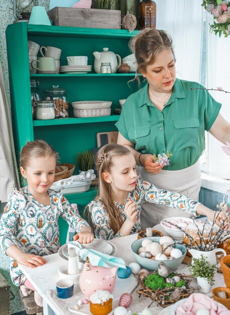 Meisjes zusters moeder aan tafel met paasdecoratie Viering in keukenTablescape voor thuis PaasvakantieFamilie religieuze traditionele feestelijke voedselmaaltijd Gekleurde eierentaart leuke konijnenoren