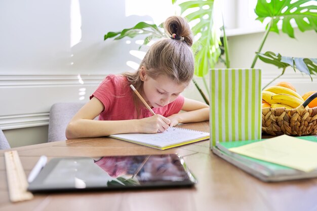 Meisjeskind dat thuis studeert, aan tafel zit. Schoolmeisje schrijft in notitieboekje, kijkt naar boek, afstandsonderwijs, moderne school en onderwijs