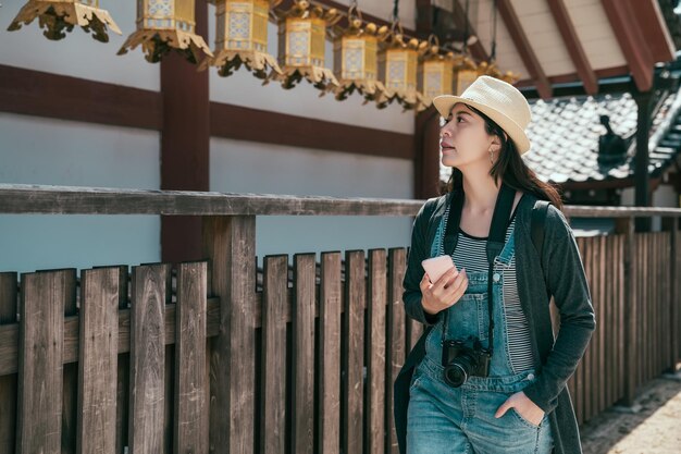 Meisjesreiziger houdt mobiele telefoon vast om online gids te lezen tijdens een bezoek aan shitennoji osaka, japan. vrouw toerist draagt camera en wandelpas houten hek. dame die op zoek is naar een gouden lantaarn die op het dak hangt
