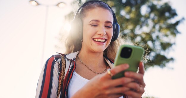 Meisjestelefoon en koptelefoon buiten voor muziekcommunicatie en sociale media met glimlach en streaming Vrouwelijke persoon smartphone en wandelen in de stad voor netwerken reizen en mobiele app met radio