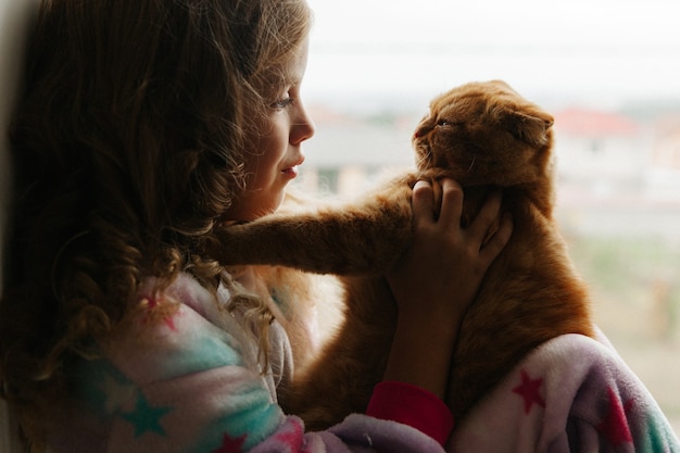 Foto meisjestiener zit in pyjama bij het raam van het huis en houdt een gemberkat vast. blijf thuis.