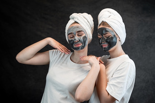 Meisjesvrienden in witte handdoek die zwart modderklei-gezichtsmasker aanbrengen dat op zwart wordt geïsoleerd