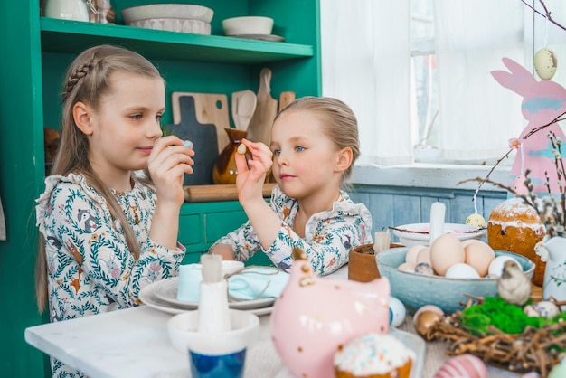 Meisjeszusjes aan tafel met paasdecoratie Feest in de keuken Tafellandschap voor thuis Paasvakantie Familie religieuze traditionele feestelijke maaltijd Gekleurde eierkoek grappig konijntje snoep zoet