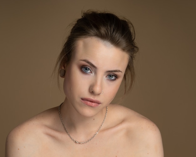 Melancholy girl with heterochromia, nose piercing and plug in one ear, and strange hairstyle. With amazing professional makeup and silver chain around her neck. Beige background. Studio shot