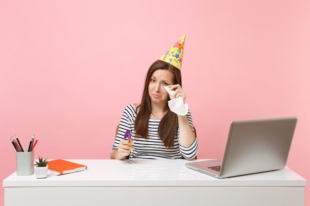 Меланхоличная женщина в партийной шляпе плачет, вытирая слезы тканью, потому что празднует день рождения в одиночестве на работе за белым столом с ноутбуком, изолированным на розовом фоне. Достижение деловой карьеры. Скопируйте пространство.