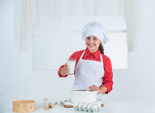 Melk als ingrediënt. kind in chef-kok uniform en hoed. tienermeisje dat deeg voorbereidt. taart maken volgens recept. tijd om te eten. gelukkig kind koken in de keuken. koekjes bakken in de keuken. professionele en bekwame bakker.