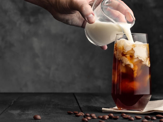 Melk wordt in een longdrinkglas met ijs en koffie op een grijze houten tafel uit een melkkan gegoten. Koffie met melk en ijs.