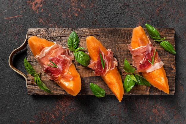 Melon cantaloupe slices with prosciutto ham and basil on black background. Italian appetizer. top view. Healthy food