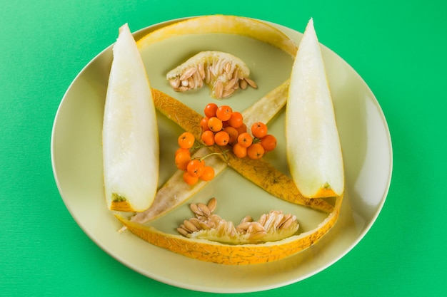 Photo melon slices melon seeds and skins on a green plate melon and rowan berries on green background vegetarian healthy food top view copy space