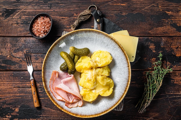 Melted Raclette Swiss cheese with boiled potato and ham. Wooden background. Top view.