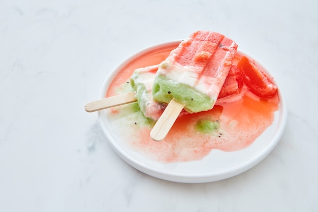 Melting fruit ice cream on a stick with splashes in a plate