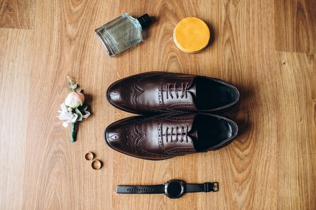 Accessori da uomo, profumo, boutonniere, anelli d'oro, orologi e scarpe di cuoio dello sposo su un pavimento di legno.