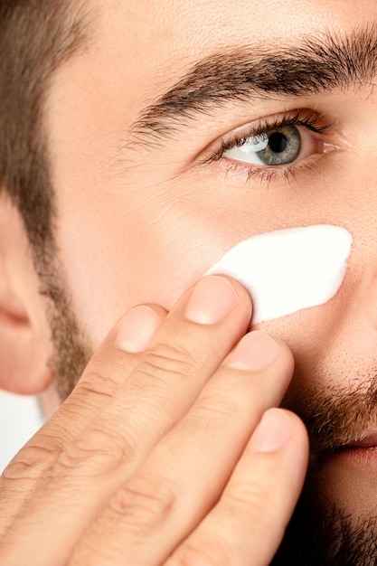 Men's beauty - Young man is applying moisturizing and anti aging cream on his face against white background