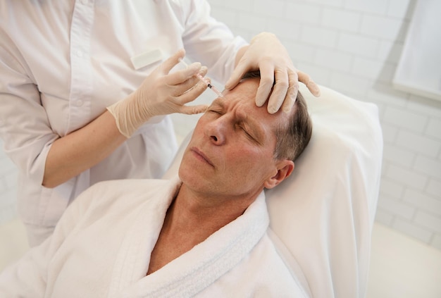 Men's cosmetology. Beautician injects a botulinum toxin into a man's forehead to relax the muscles. Mature man receiving facial rejuvenation injection procedure for smoothing and anti-aging treatment