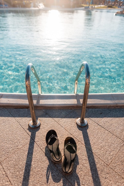 男性用のビーチサンダルの靴は、日没時に青い水でプールの近くに立っています。夏休みのコンセプト