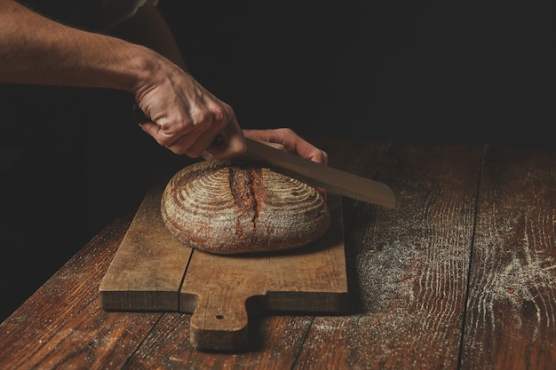 Men's hands cut round rye bread on a wooden brown cutting board