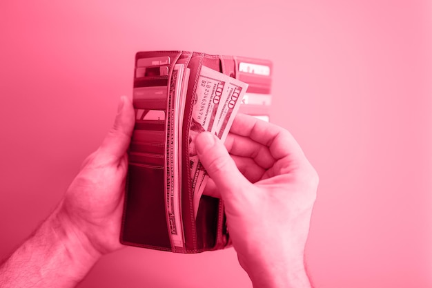 Men's hands hold 100 dollar bills and a wallet on a pink background toned in viva magenta trend color of the year 2023