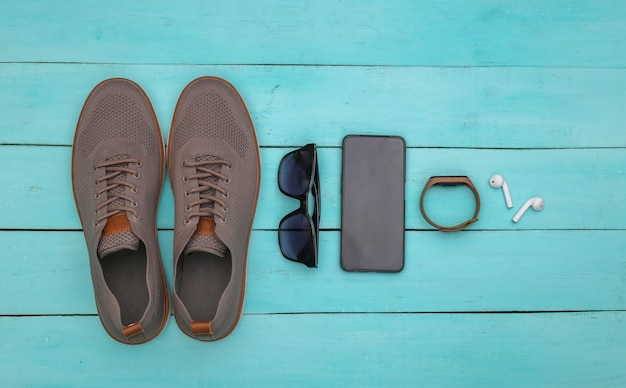 Men's shoes and gadgets (smartphone, smart bracelet, headphones), sunglasses on a blue wooden background. Top view