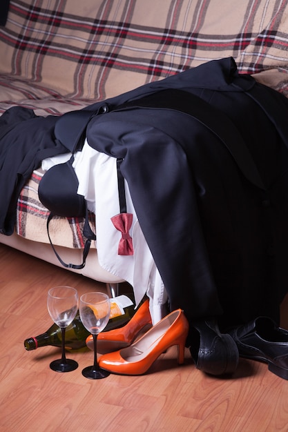 Men's, women's clothes and shoes are scattered on the couch after the party. An empty wine bottle and glasses stand on the floor