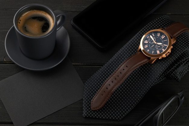 Men's wrist watch on a black tie, a hot cup of coffee and a smartphone on a wooden table. black envelope with place for text.