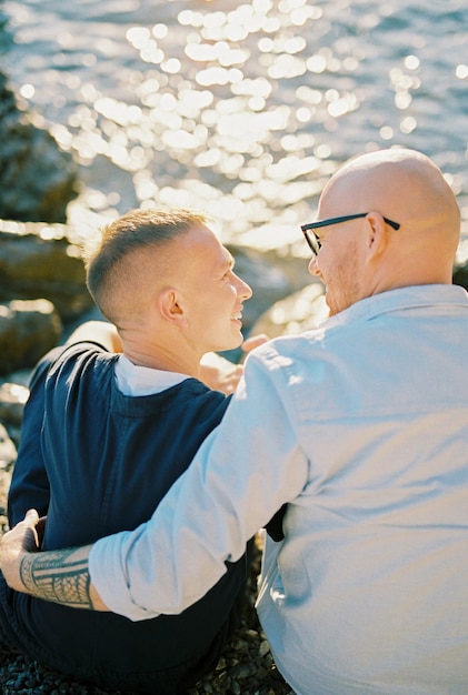 Photo men sit hugging on a pebble beach and look at each other back view