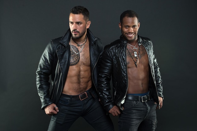 Men on smiling faces with bristle and tattoos. Fashion and style concept. Men with sexy muscular torsos look brutally. Machos with muscular torsos look attractive in leather jackets, dark background.