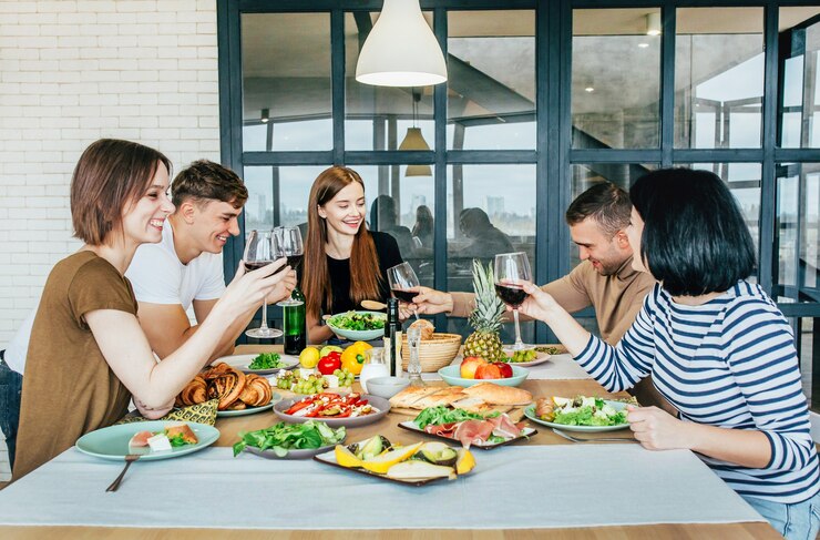 Men and women in the kitchen are sitting at a laid table with glasses of wine in their hands laughing and talking