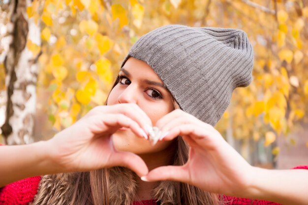 Mening van jonge vrouw met grijze wollen hoed die een hartvorm met haar vingers in het park maakt
