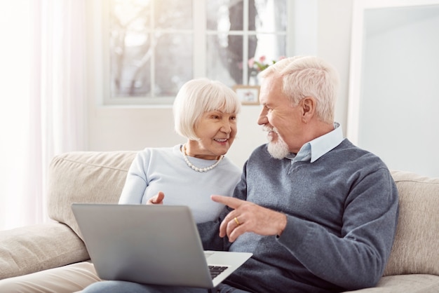 Meningen delen. Vrolijke senior man en vrouw zittend op de bank in de woonkamer en samen een artikel bespreken, het online hebben gelezen