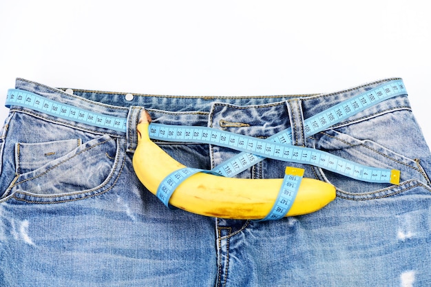 Mens denim broek met banaan imiteren mannelijke geslachtsorganen Gezondheid en mannelijke seksualiteit concept Kinky fruit op denim stof close-up Blue jeans met meetlint in plaats van riem op witte achtergrond