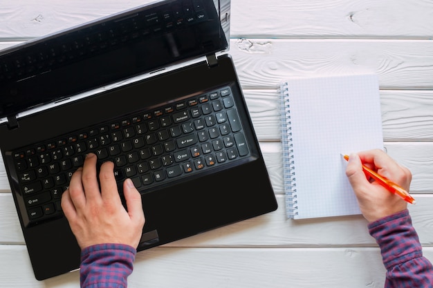 Mens die bij de computer werkt en in een notitieboekje op een witte houten achtergrond schrijft