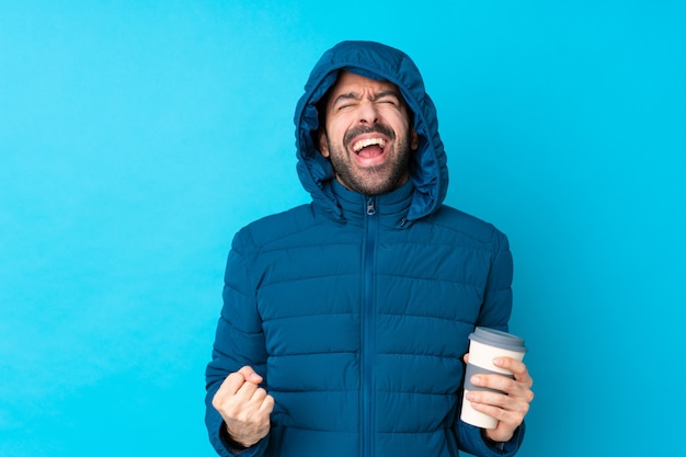 Mens die de winterjasje draagt en een meeneemkoffie over geïsoleerde blauwe muur houdt die aan de voorzijde met wijd open mond schreeuwt