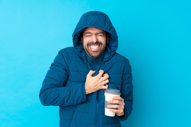 Mens die de winterjasje draagt en een meeneemkoffie over geïsoleerde blauwe muur houdt die een pijn in het hart heeft