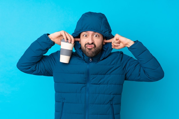 Foto mens die de winterjasje draagt en een meeneemkoffie over geïsoleerde blauwe muur houdt gefrustreerd en het behandelen van oren