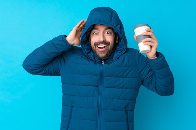 Mens die de winterjasje draagt en een meeneemkoffie over geïsoleerde blauwe muur met verrassingsuitdrukking houdt