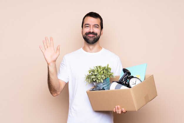 Mens die een doos houdt en zich in nieuw huis beweegt die met hand met gelukkige uitdrukking groeten