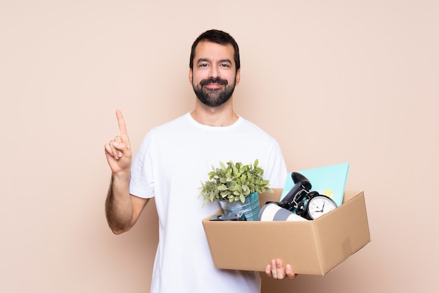 Mens die een doos houdt en zich in nieuw huis over het geïsoleerde tonen beweegt en een vinger in teken van het beste opheft