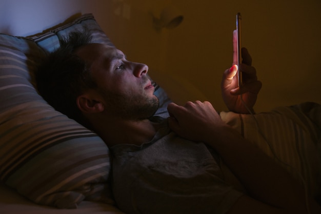 Mens die in bed liggen die smartphone gebruiken bij nacht. Slapeloosheid, melatonine, verslaving aan sociale netwerken