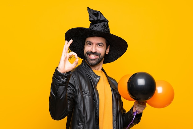 Mens die met heksenhoed zwarte en oranje luchtballons houden voor Halloween-partij die ok teken met vingers tonen