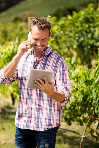 Mens die op telefoon spreekt terwijl het gebruiken van tablet bij wijngaard