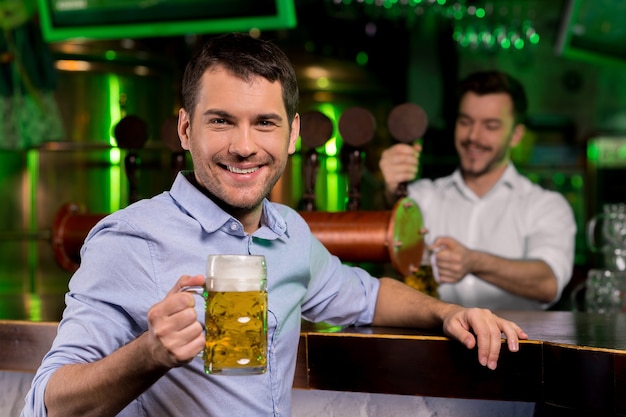 Mens in bierkroeg. Knappe jonge man die een bierpul vasthoudt en glimlacht terwijl de barman bier op de achtergrond schenkt