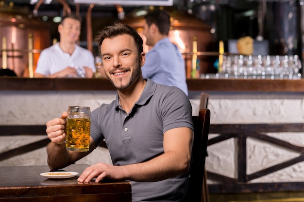 Mens in bierkroeg. Vrolijke jonge man die een bierpul vasthoudt en glimlacht terwijl hij in de bar zit