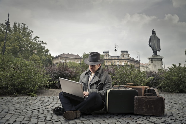 Mens met bagage die op de straat situeert en laptop met behulp van