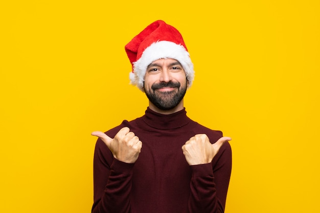 Mens met Kerstmishoed over geïsoleerde gele muur met duimen op gebaar en het glimlachen