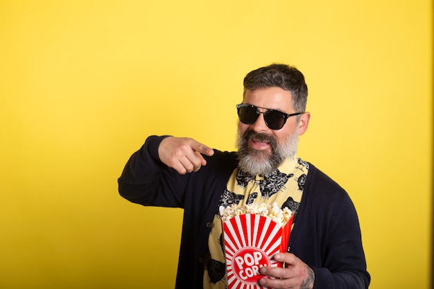 Mens met witte baard en zonnebril op geïsoleerde gele achtergrond die een grote emmer popcorn houden terwijl vooruit het richten