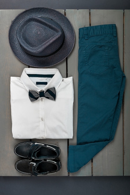 Mens outfit on wooden background, Kids fashion clothes, Grey fedora, navy pant, white shirt, black bow tie and boat shoes for boy, top view, flat lay, copy space.