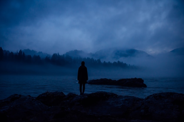 Menselijk silhouet in een dikke blauwe mist op de achtergrond van beboste heuvels en bergrivier. eenzame vrouwenfiguur op een mysterieuze kust. contemplatie, meditatie, eenheid in de natuur.