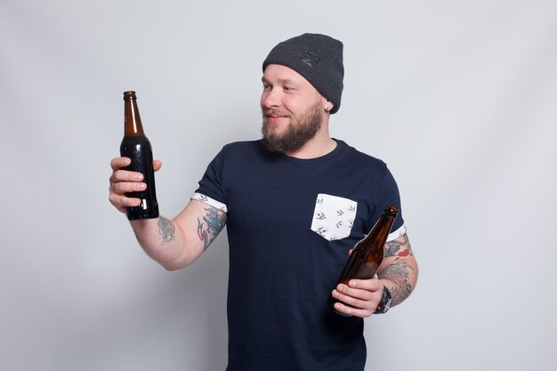 Mensen, 4K en lifestyle concept - brutale bebaarde man met getatoeëerde arm drinkt een biertje uit een fles. knappe man met hoed. Brutale bebaarde jongen met tattoo