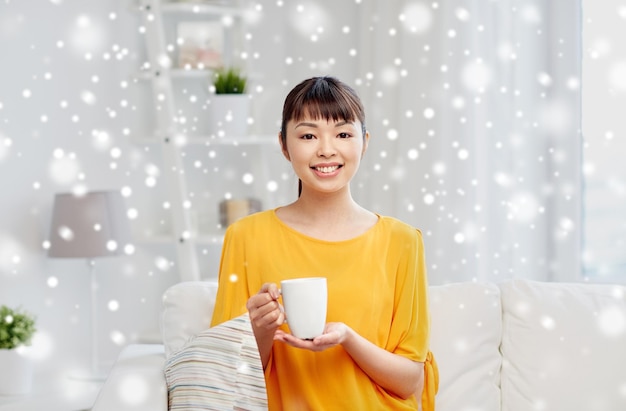 mensen, dranken en vrijetijdsconcept - gelukkige Aziatische vrouw zit op de bank en drinkt thee uit een kopje of beker thuis over sneeuw