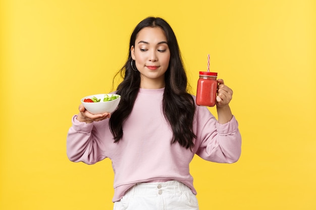 Mensen emoties, gezonde levensstijl en voedsel concept. Schattig glimlachend Aziatisch meisje dat denkt aan salade met een tevreden grijns, smoothie vasthoudt, gele achtergrond.